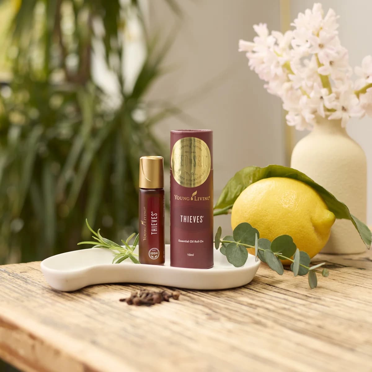 Young Living Thieves Roll-On 10 ml steht mit seiner Verpackung auf einer weißen Schale, daneben Zitrone, Rosmarin, Eukalyptus und Nelken auf einem Holztisch.