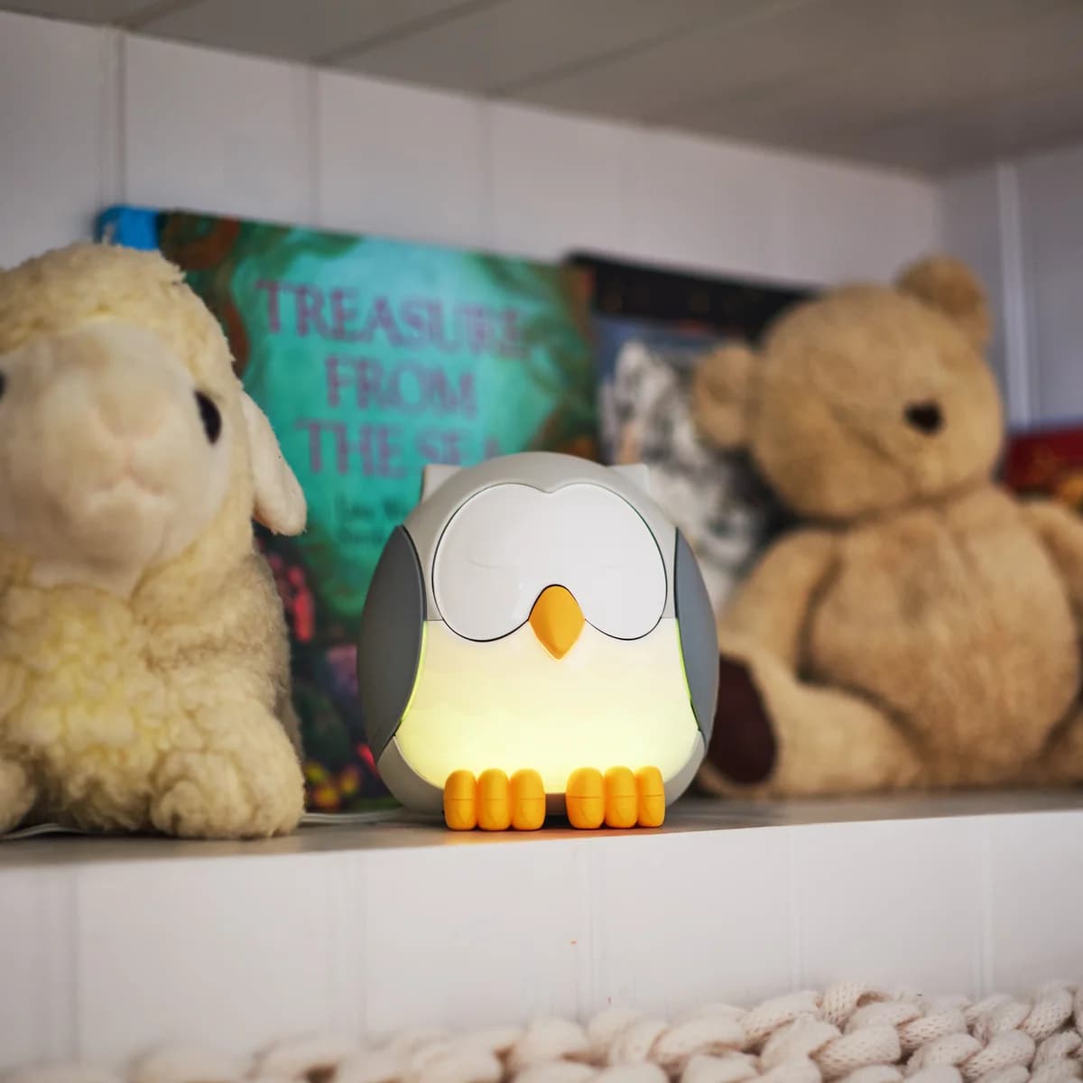 Der Young Living Feather The Owl Ultrasonic Diffuser leuchtet warm auf einem weißen Regal zwischen Plüschschaf und Teddybär, dahinter Kinderbücher.