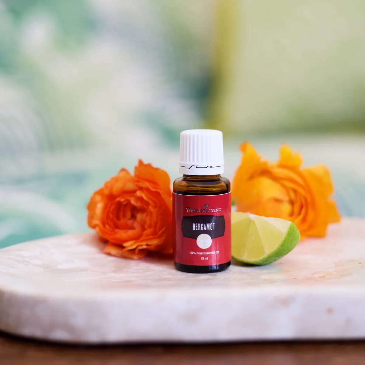 Ein Fläschchen Young Living Bergamottenöl 15 ml steht auf einer hellen Steinplatte zwischen zwei orangefarbenen Blüten und einem Limettenstück.