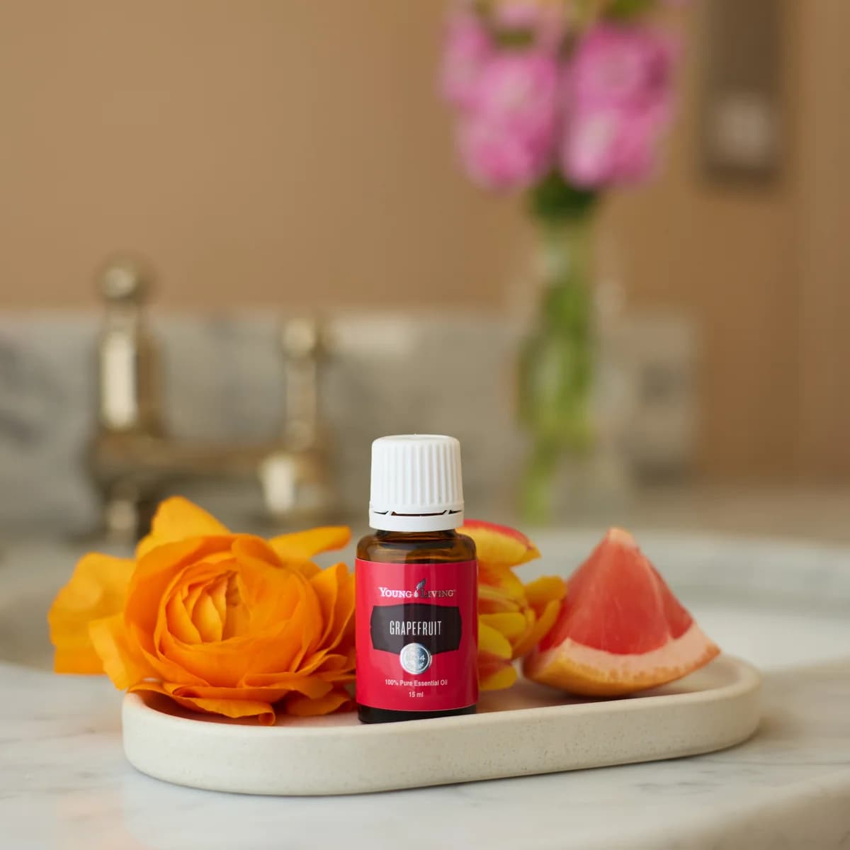 Ein Fläschchen Young Living Grapefruitöl 15 ml steht auf einem ovalen Keramiktablett zwischen orangefarbenen Blüten und einem Grapefruitstück am Waschtisch.