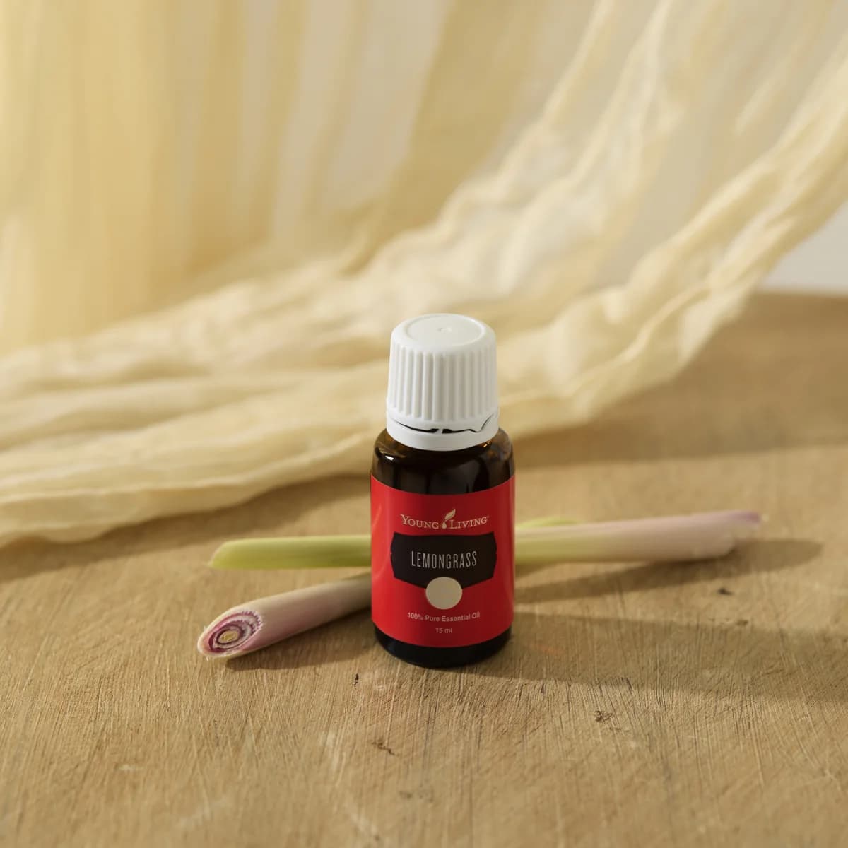 Ein Fläschchen Young Living Zitronengrasöl 15 ml steht auf einem Holztisch vor zwei Zitronengrasstängeln, im Hintergrund ein drapiertes helles Tuch.