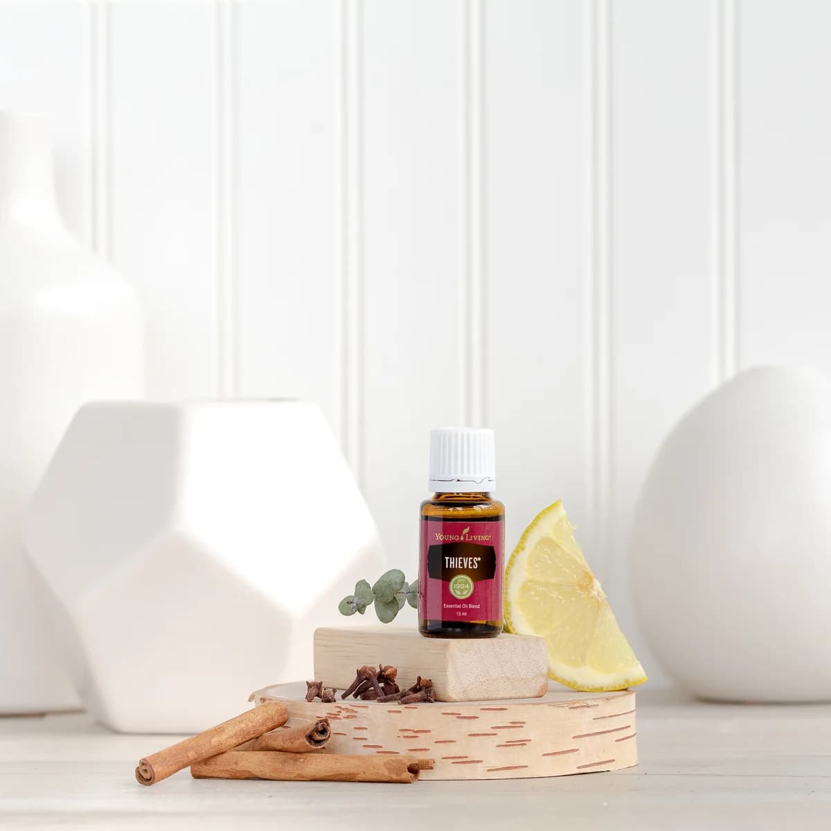 Ein Fläschchen Young Living Thieves 15 ml steht auf einem Holzklotz zwischen Nelken, Eukalyptus und einer Zitronenscheibe vor weißen Keramikvasen.