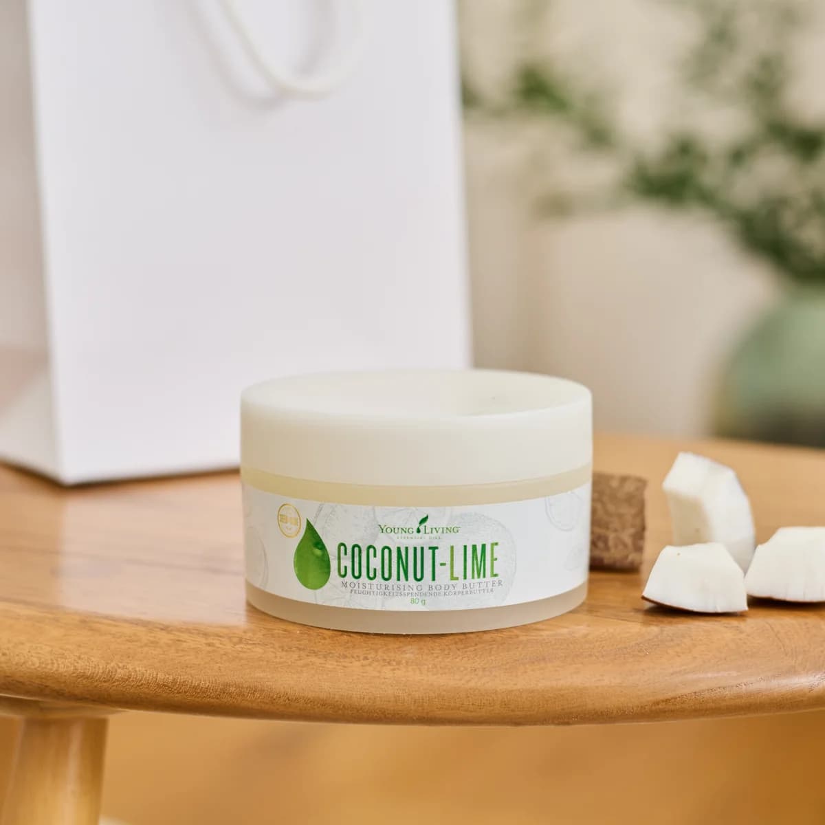 Ein Tiegel Young Living Coconut-Lime Moisturising Body Butter 80 g steht auf einem runden Holztisch neben Kokosstücken und einer weißen Papiertüte.