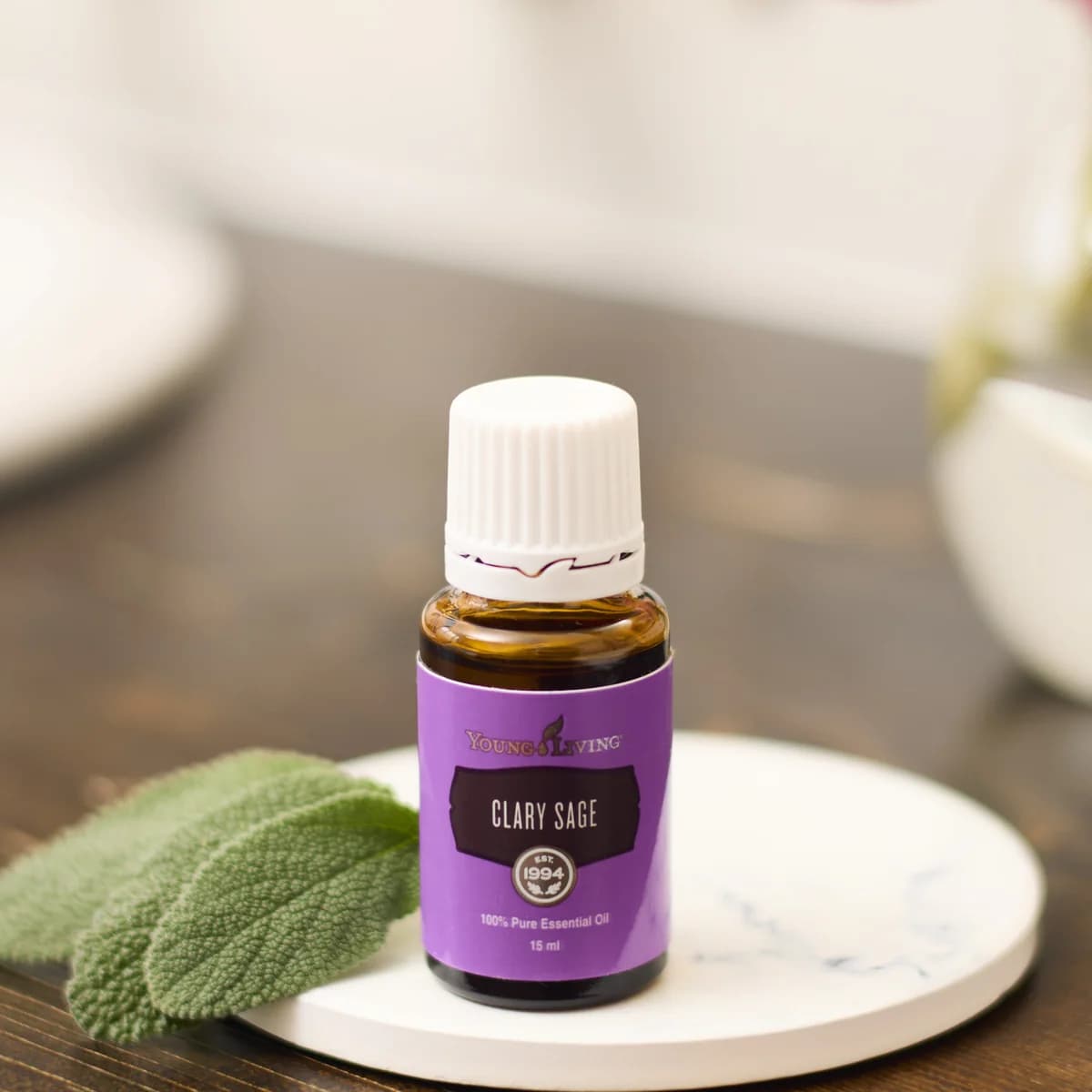 Ein Fläschchen Young Living Muskatellersalbeiöl 15 ml steht auf einem weißen Marmoruntersetzer neben Salbeiblättern, dahinter rosa Tulpen.