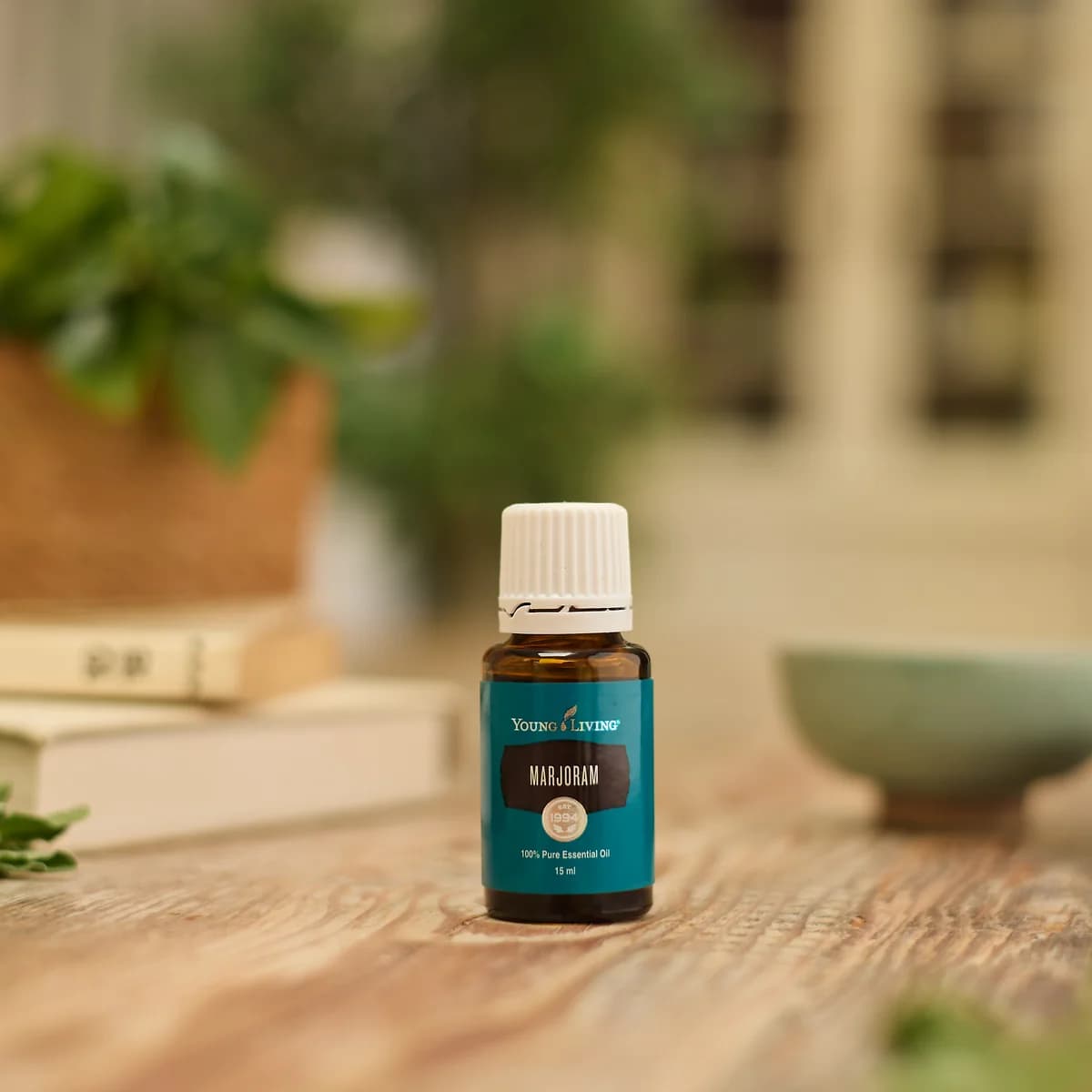 Ein Fläschchen Young Living Majoranöl 15 ml steht auf einem Holztisch, dahinter unscharf eine Topfpflanze und ein grünes Schälchen.