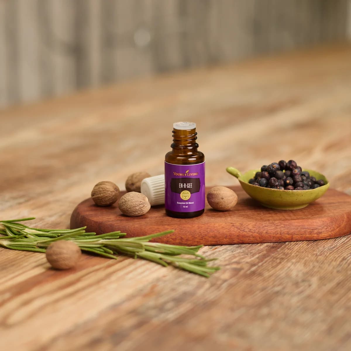 Ein geöffnetes Fläschchen Young Living En-R-Gee 15 ml steht auf einem Holzbrett neben Muskatnüssen, Rosmarinzweigen und einer Schale Wacholderbeeren.