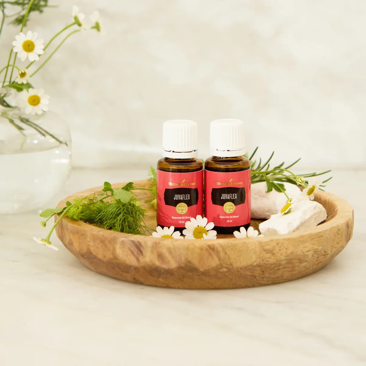 Zwei Fläschchen Young Living JuvaFlex 15 ml stehen in einer Holzschale mit Kamillenblüten, Rosmarin und hellen Steinen auf einer Marmorfläche.