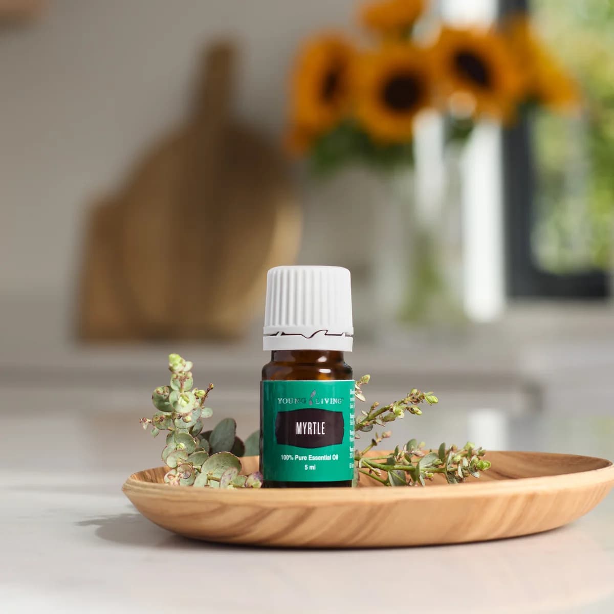 Ein Fläschchen Young Living Myrtenöl 5 ml steht auf einem Holzteller zwischen grünen Myrtenzweigen, im Hintergrund eine Küche mit Sonnenblumen.