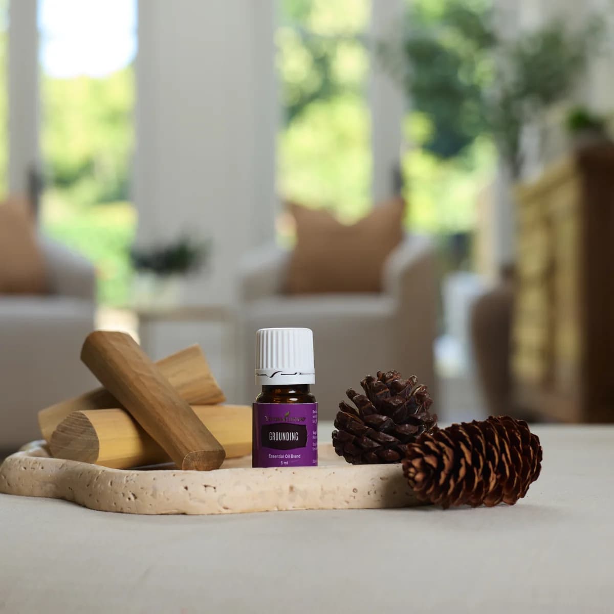 Ein Fläschchen Young Living Grounding 5 ml steht auf einem Travertintablett zwischen Holzstücken und zwei Tannenzapfen in einem hellen Wohnzimmer.