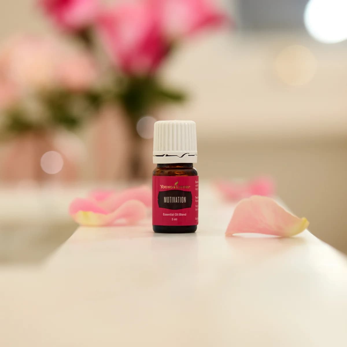 Ein Fläschchen Young Living Motivation 5 ml steht auf einer weißen Fläche zwischen rosa Rosenblättern, im Hintergrund unscharfe rosa Rosen.