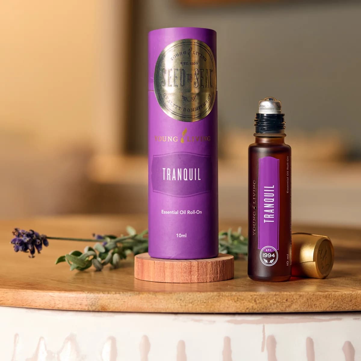 Ein Young Living Tranquil Roll-On 10 ml steht geöffnet neben seiner violetten Hülle und einem Lavendelzweig auf einem runden Holzbrett.