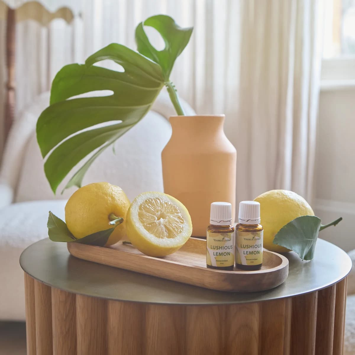 Zwei Fläschchen Young Living Lushious Lemon 15 ml stehen auf einem ovalen Holztablett neben Zitronen und einer Vase vor einem hellen Sessel.