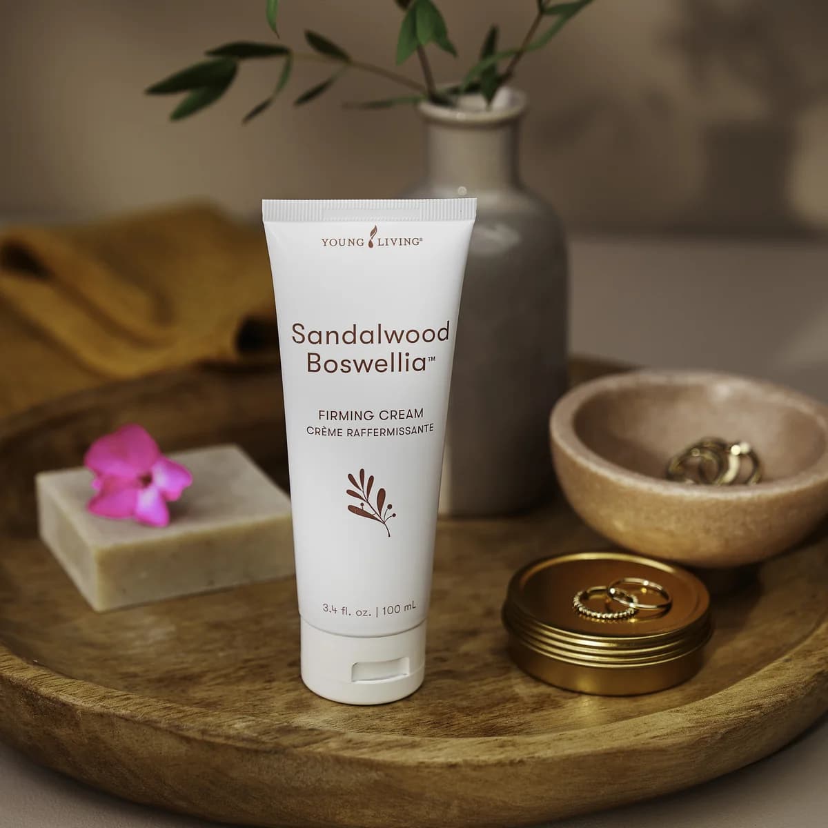 Eine Tube Young Living Sandalwood Boswellia Firming Cream 100 ml steht auf einem runden Holztablett neben Seife, einer Vase und Schalen mit Ringen.