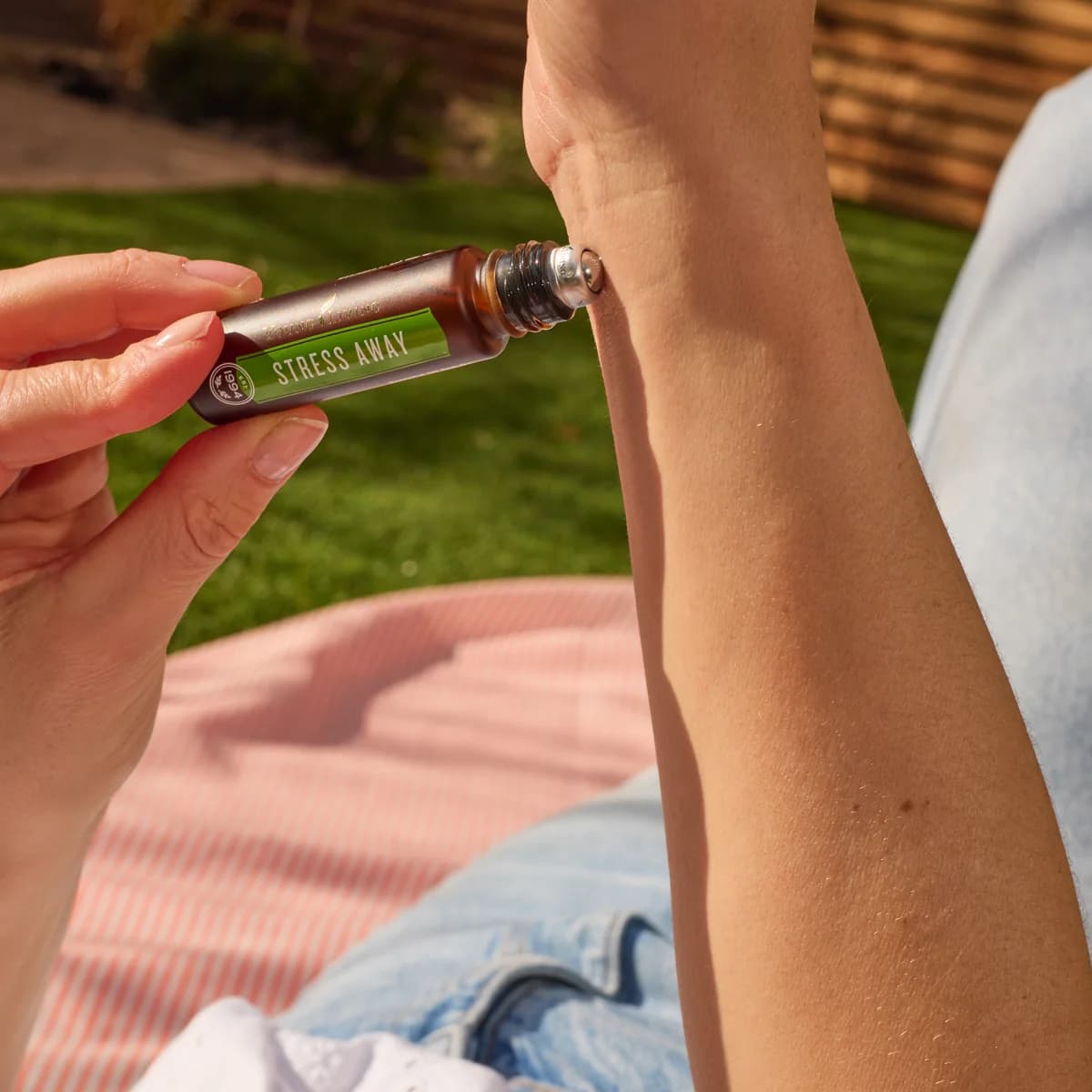 Eine Hand rollt den Young Living Stress Away Roll-On 10 ml auf die Innenseite eines Handgelenks, im Hintergrund Rasen und eine rosa gestreifte Decke.