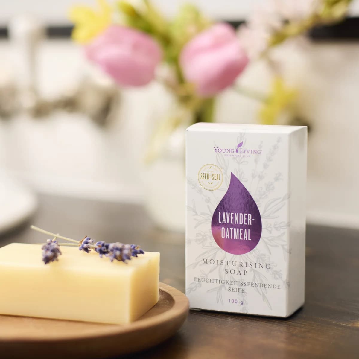 Die Schachtel Young Living Bar Soap Lavender Oatmeal steht auf dunklem Holz neben einem Seifenstück mit Lavendelblüten auf einer Holzschale, dahinter Tulpen.