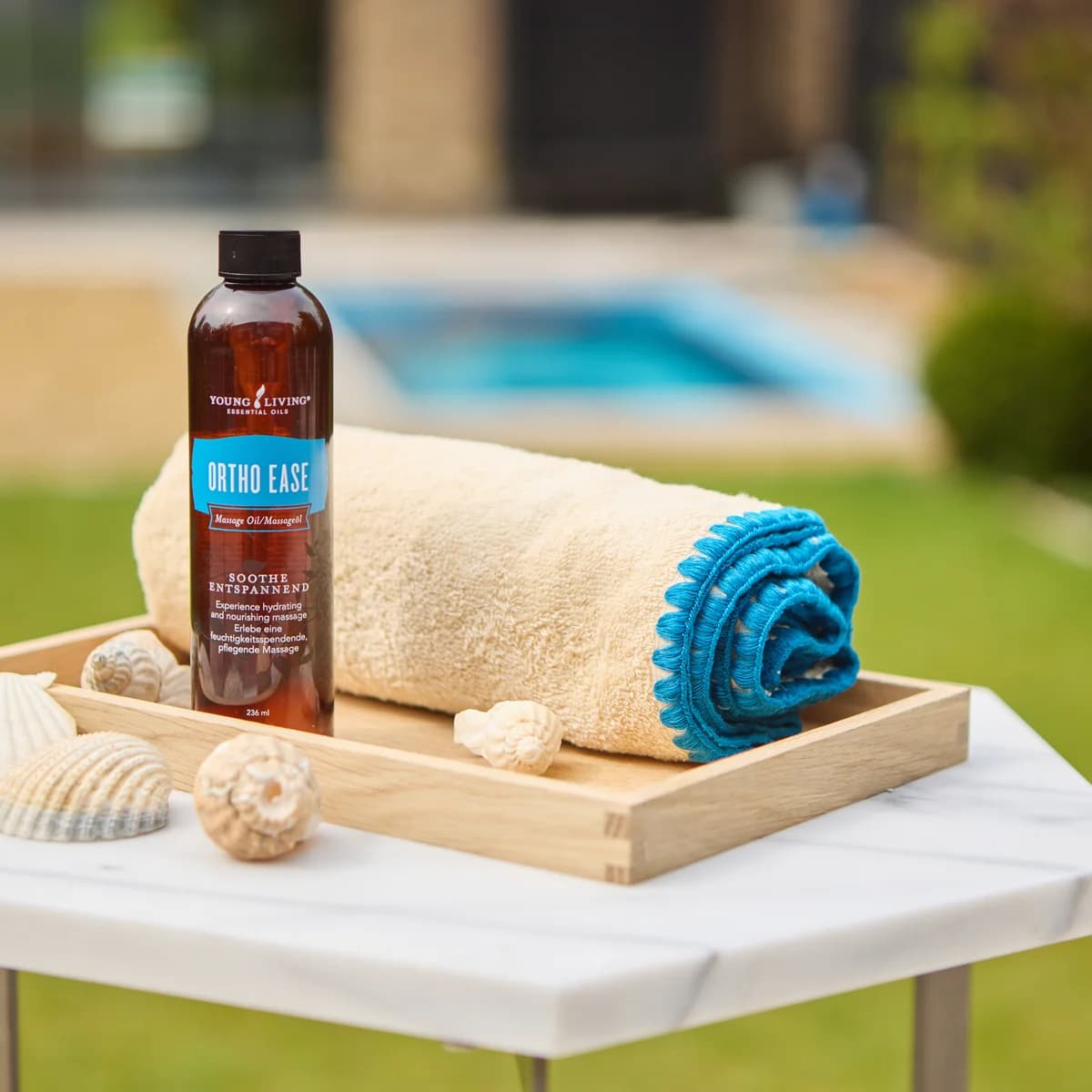 Eine Flasche Young Living Ortho Ease Massage Oil 236 ml steht auf einem Holztablett neben einem gerollten Handtuch und Muscheln, dahinter ein Pool.