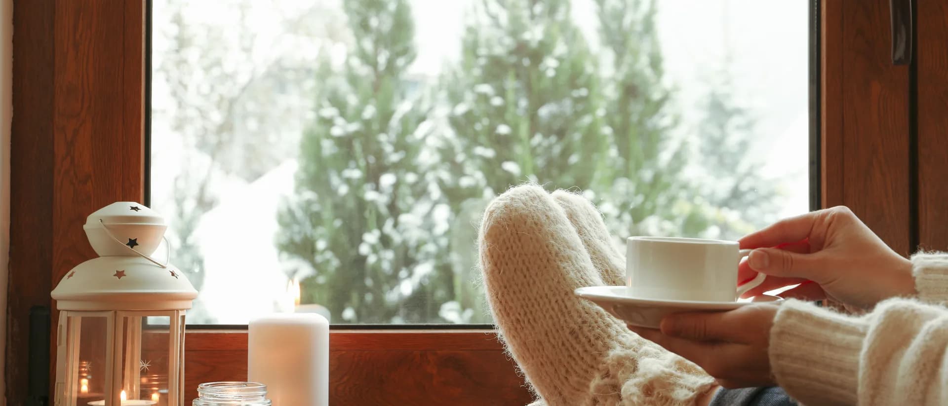 Füße in Wollsocken und eine Tasse Tee auf der Fensterbank, davor Kerzenlicht und der Blick auf verschneite Bäume vor dem Fenster