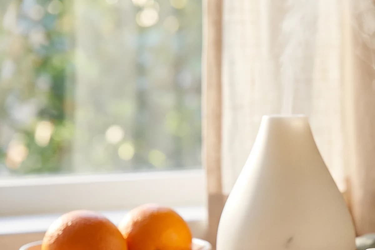 Ein weißer Diffuser mit aufsteigendem Nebel vor einem Fenster mit grünem Bokeh und einem Leinenvorhang, im Vordergrund unscharf zwei Orangen