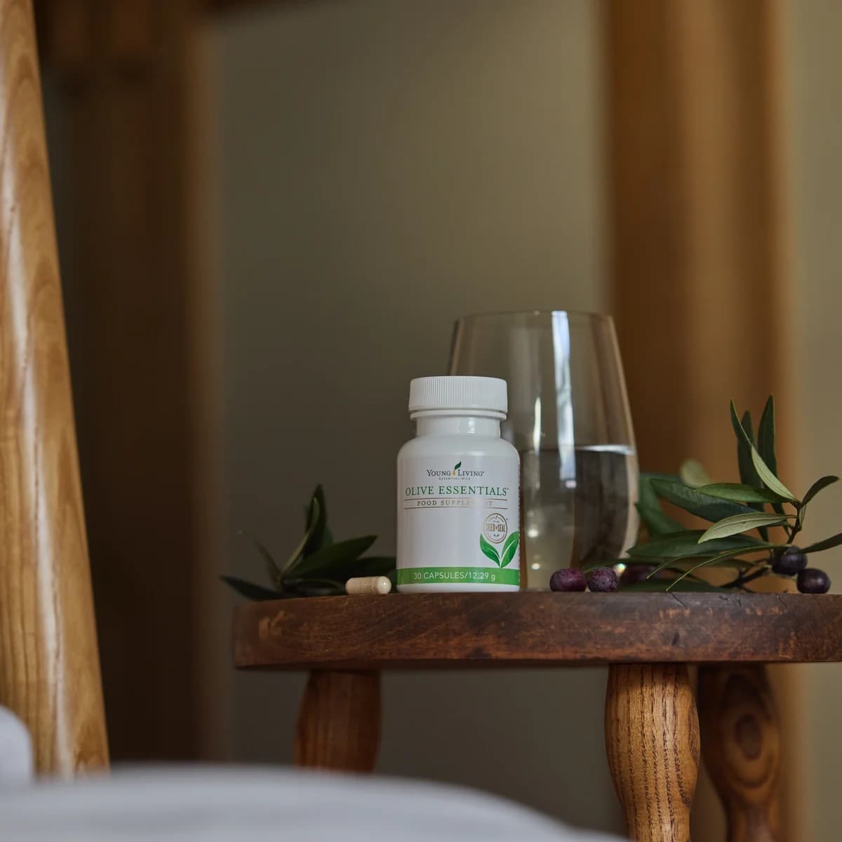 Eine Kapseldose Young Living Olive Essentials steht auf einem kleinen Holzhocker neben einem Wasserglas, Olivenzweigen und Oliven.