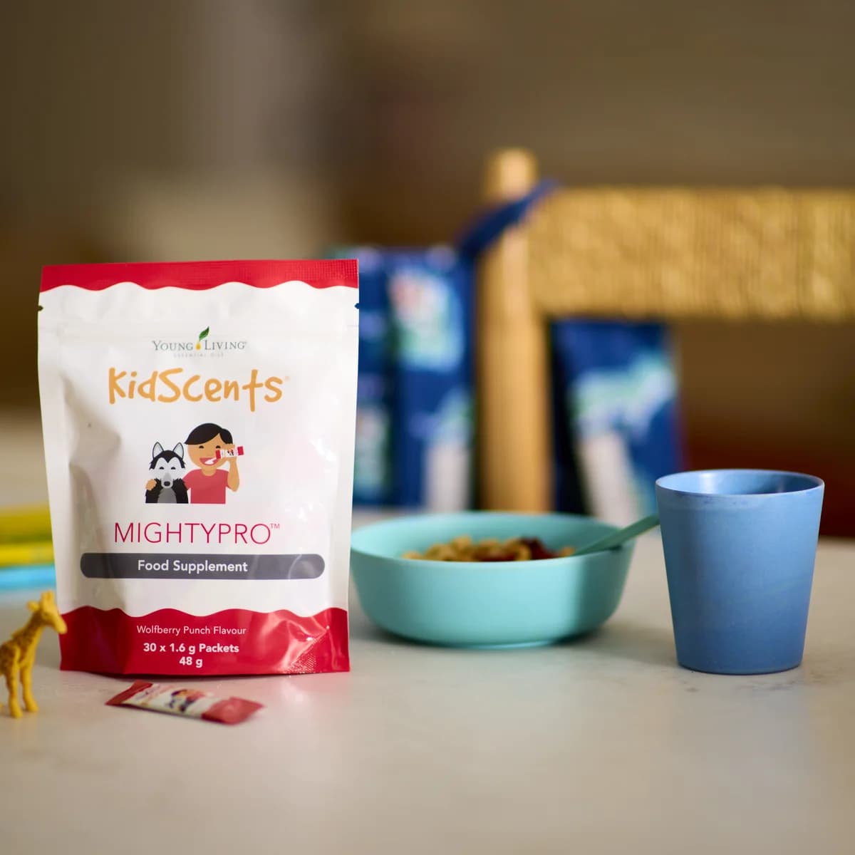 Ein Beutel Young Living KidScents MightyPro steht auf einem Tisch neben einem Portionsstick, einer Giraffenfigur, einer Müslischale und einem blauen Becher.