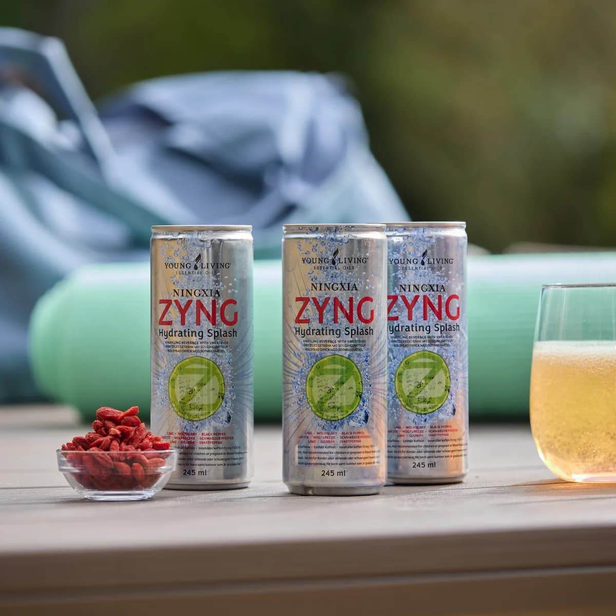 Drei Dosen Young Living NingXia Zyng stehen auf einem Gartentisch neben einem Glas mit Getränk und einer Schale Gojibeeren, dahinter eine Yogamatte.