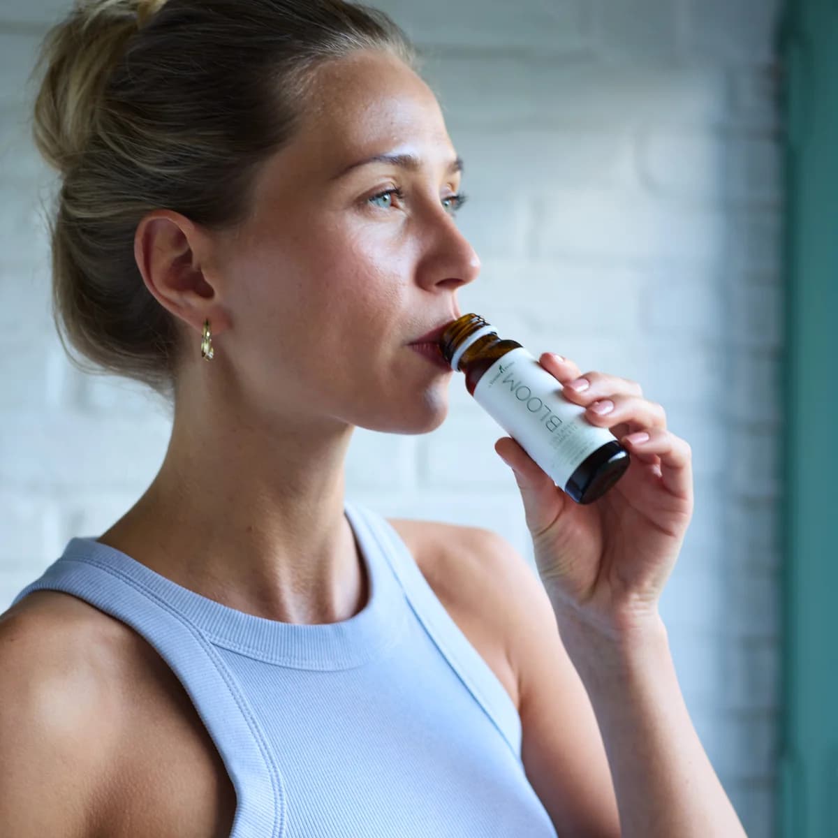 Eine Frau trinkt vor einer weißen Backsteinwand aus einem kleinen Fläschchen Young Living Bloom Collagen Complete.