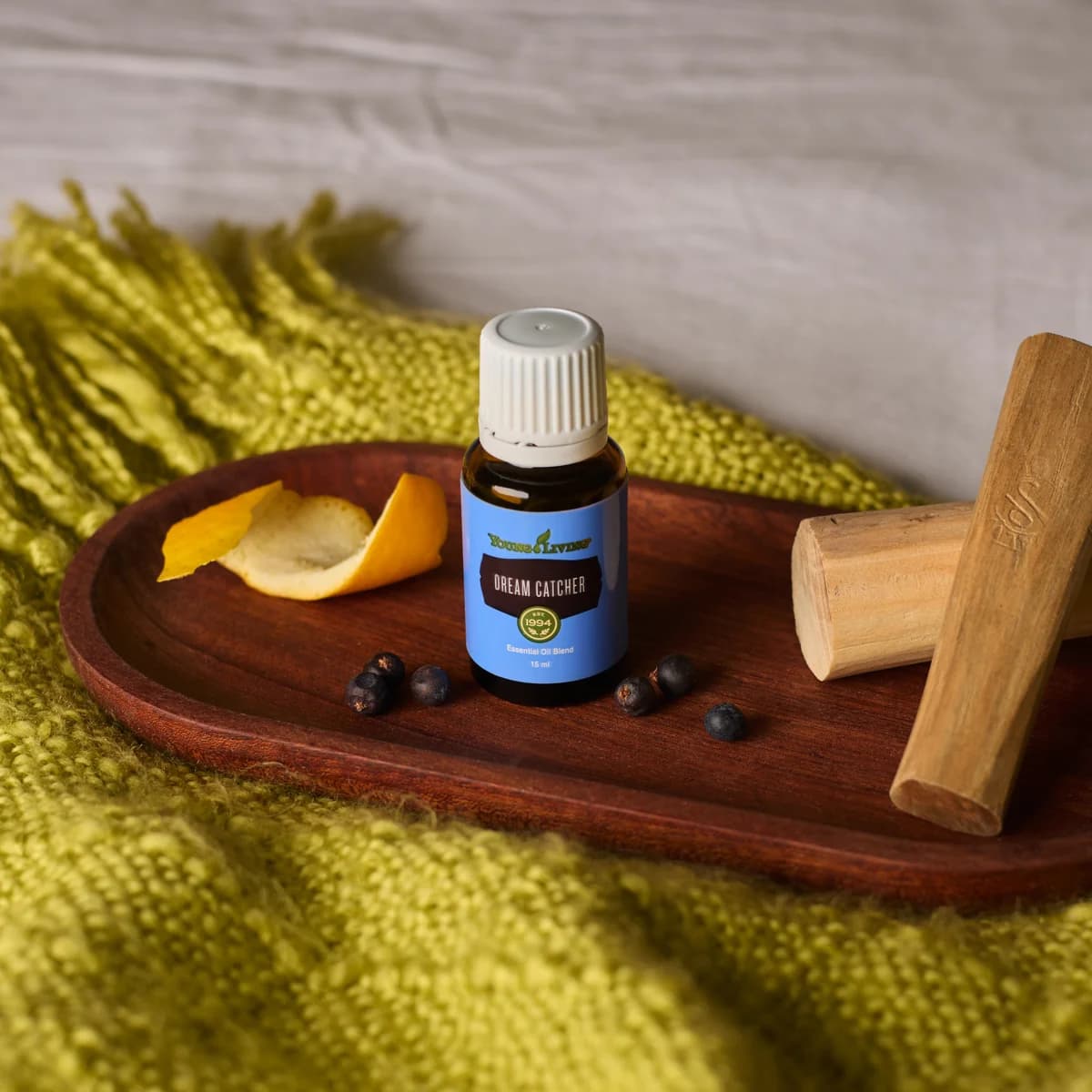 Ein Fläschchen Young Living Dream Catcher 15 ml steht auf einem dunklen Holztablett mit Orangenschale, Wacholderbeeren und Sandelholz auf einer gelben Decke.
