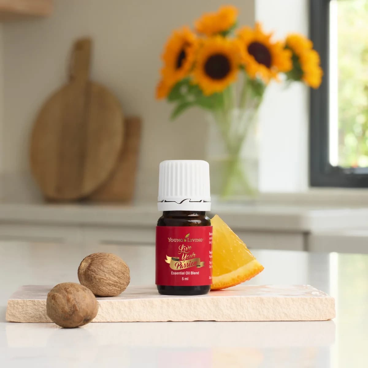 Ein Fläschchen Young Living Live Your Passion 5 ml steht auf einem hellen Steinbrett neben zwei Muskatnüssen und einer Orangenspalte in einer Küche.