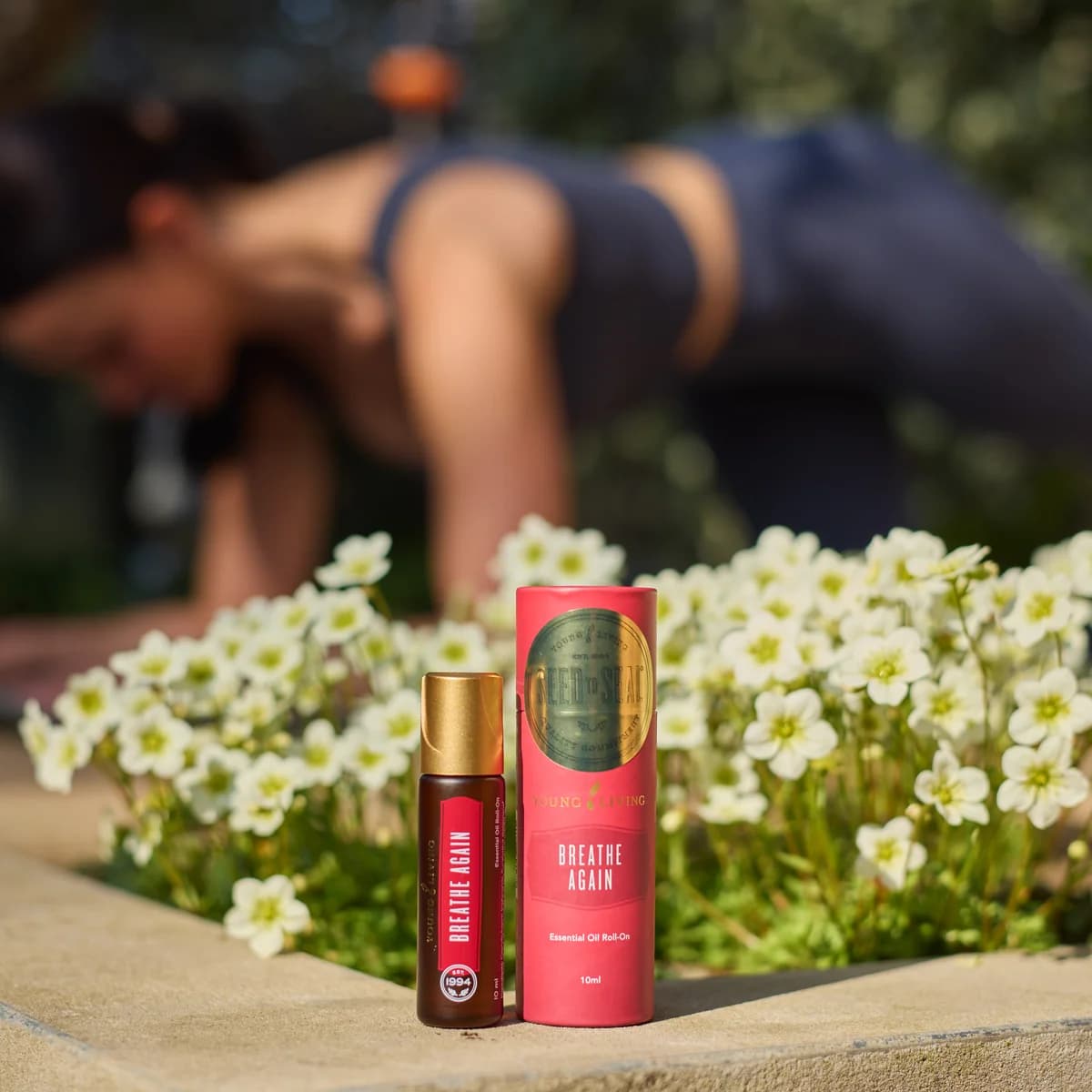 Ein Young Living Breathe Again Roll-On 10 ml steht neben seiner roten Hülle auf einer Steinkante vor weißen Blüten, dahinter trainiert eine Frau im Freien.