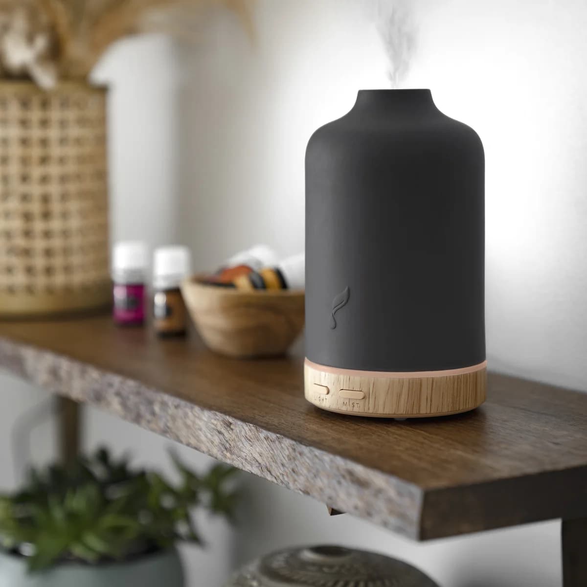 Der schwarze Young Living Ember Diffuser mit Holzsockel steht auf einem Holzregal neben zwei Ölfläschchen und einer Schale, aus der Öffnung steigt Nebel.