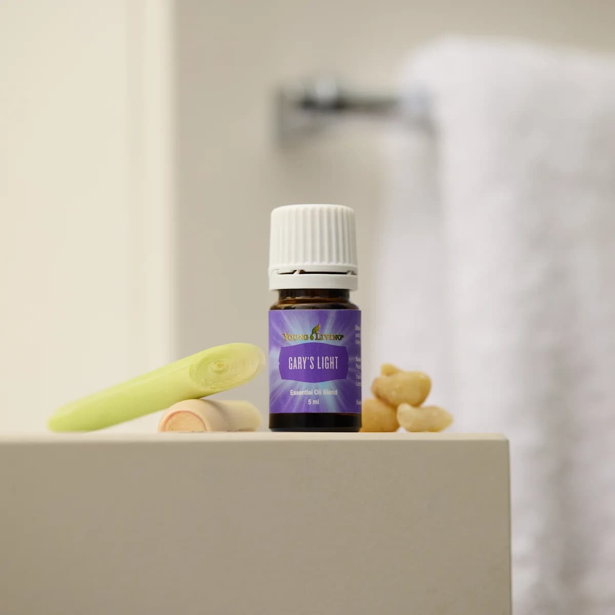 Ein Fläschchen Young Living Gary's Light 5 ml steht auf einer hellen Ablage im Bad zwischen einem Zitronengrasstängel und Harzstücken, dahinter ein Handtuch.