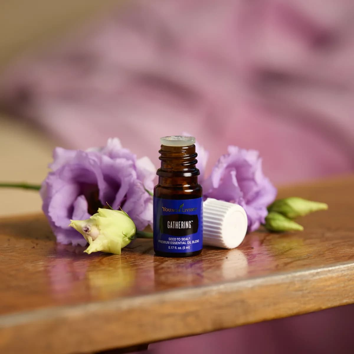 Ein geöffnetes Fläschchen Young Living Gathering 5 ml steht mit abgelegtem Deckel auf einem Holztisch zwischen lila Blüten vor violettem Stoff.