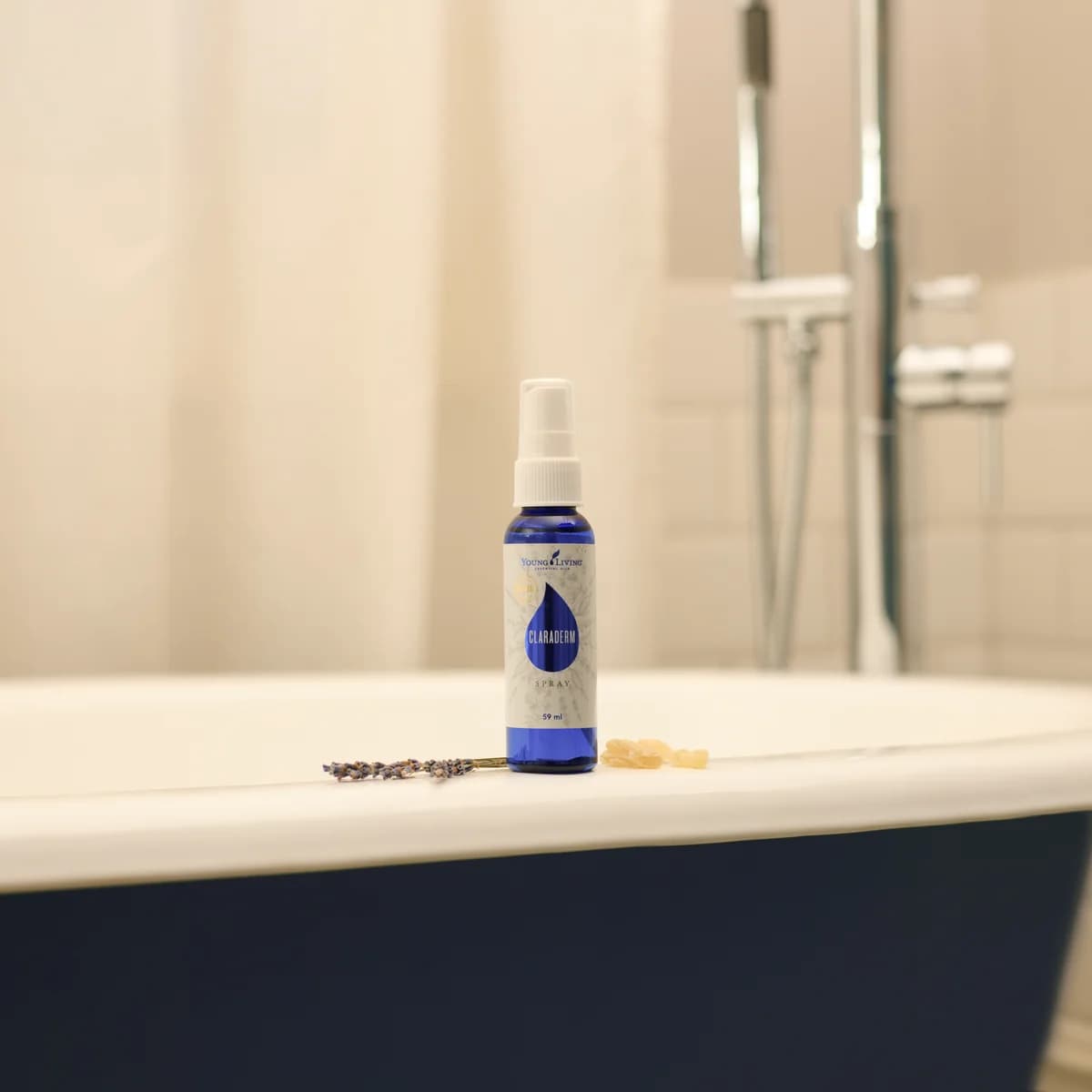 Eine blaue Sprühflasche Young Living ClaraDerm Spray steht auf dem Rand einer Badewanne neben einem Lavendelzweig und Harzstückchen, dahinter weiße Fliesen.