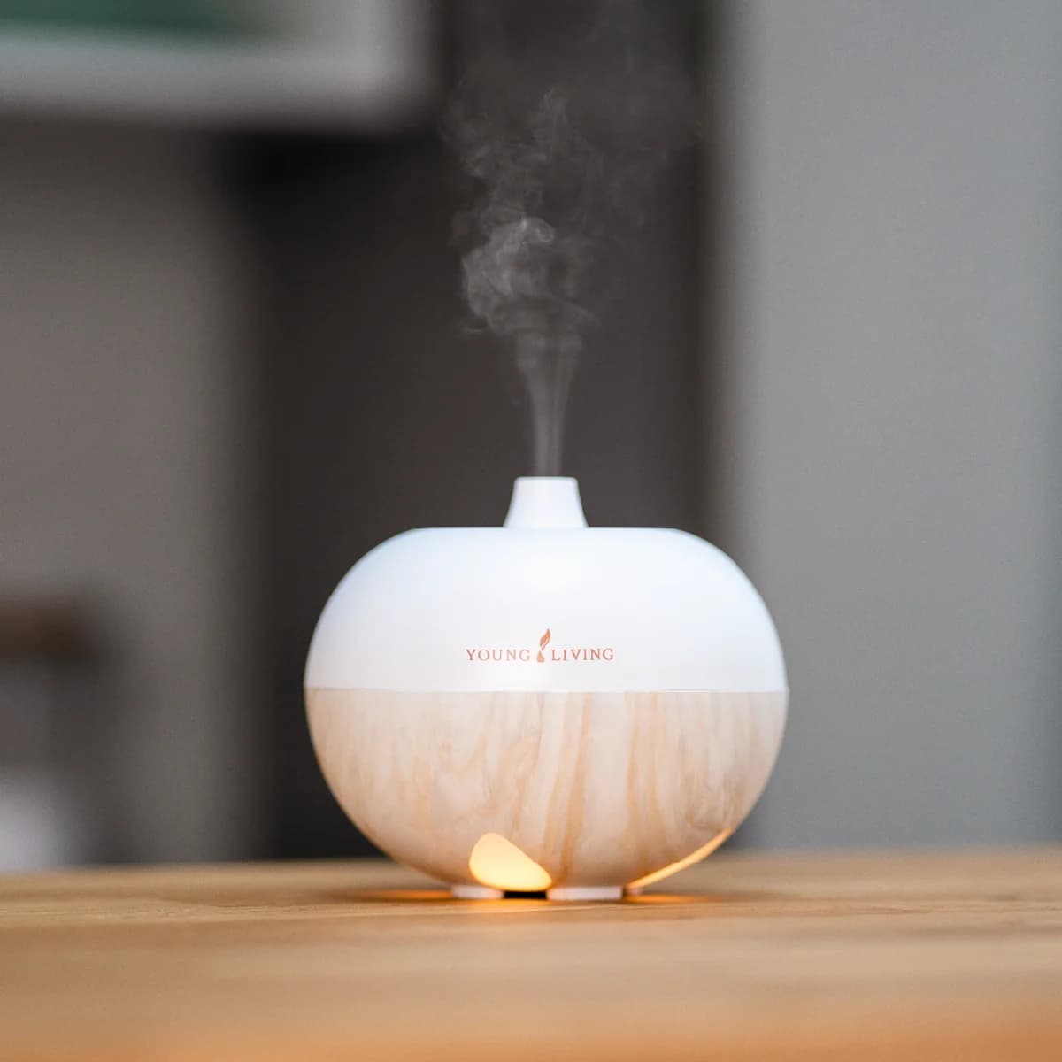 Der Young Living AromaGlobe Ultrasonic Diffuser mit Sockel in Holzoptik steht auf einem Holztisch, Nebel steigt auf und der Fuß leuchtet warm.