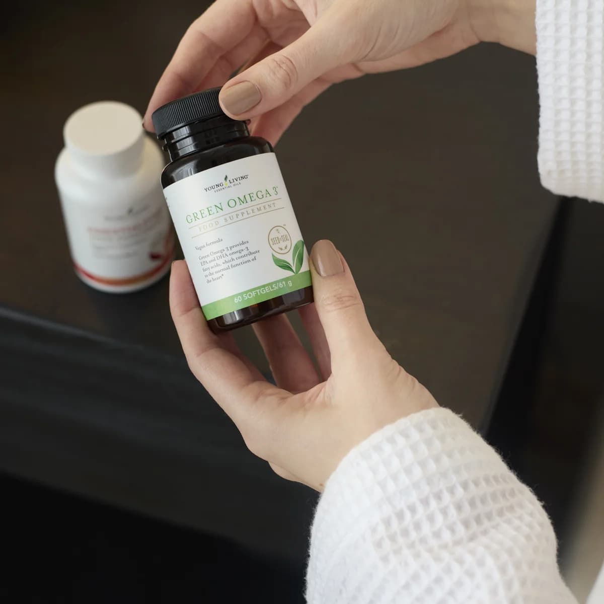 Zwei Hände im weißen Bademantel halten eine braune Dose Young Living Green Omega 3, dahinter steht eine zweite Dose auf einer dunklen Ablage.