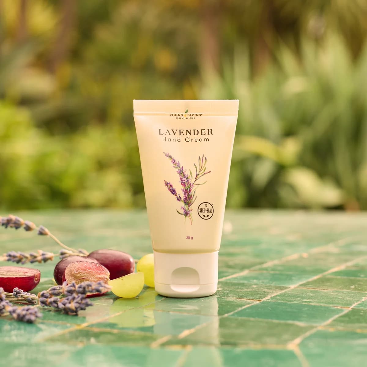 Eine Tube Young Living Lavender Hand Cream 28 g steht auf einem grün gefliesten Tisch im Freien neben Lavendelzweigen und halbierten Weintrauben.