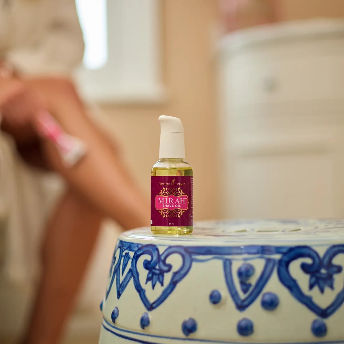 Ein Fläschchen Young Living Mirah Shave Oil 59 ml steht auf einem blau-weißen Keramikhocker, dahinter rasiert sich eine Frau im Bademantel das Bein.