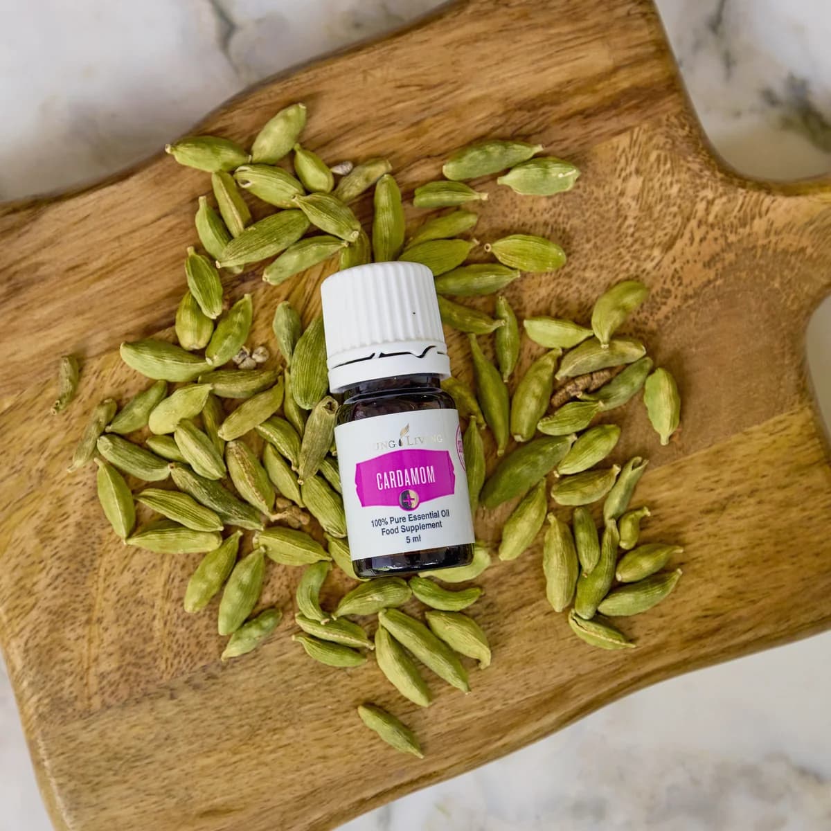 Ein Fläschchen Young Living Cardamom+ 5 ml liegt auf einem Holzbrett zwischen grünen Kardamomkapseln auf hellem Marmor.