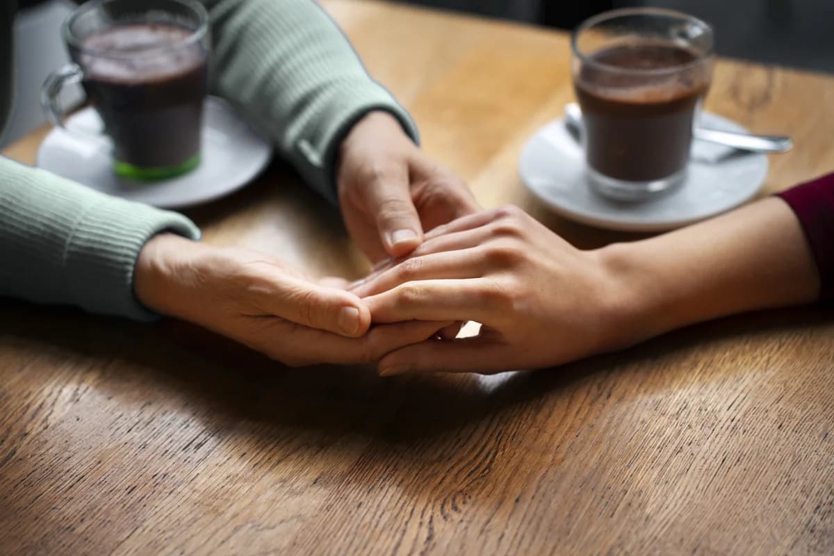 Zwei Hände auf einer Holztischplatte: eine ältere Hand hält eine jüngere, daneben zwei Gläser heiße Schokolade auf Untertassen