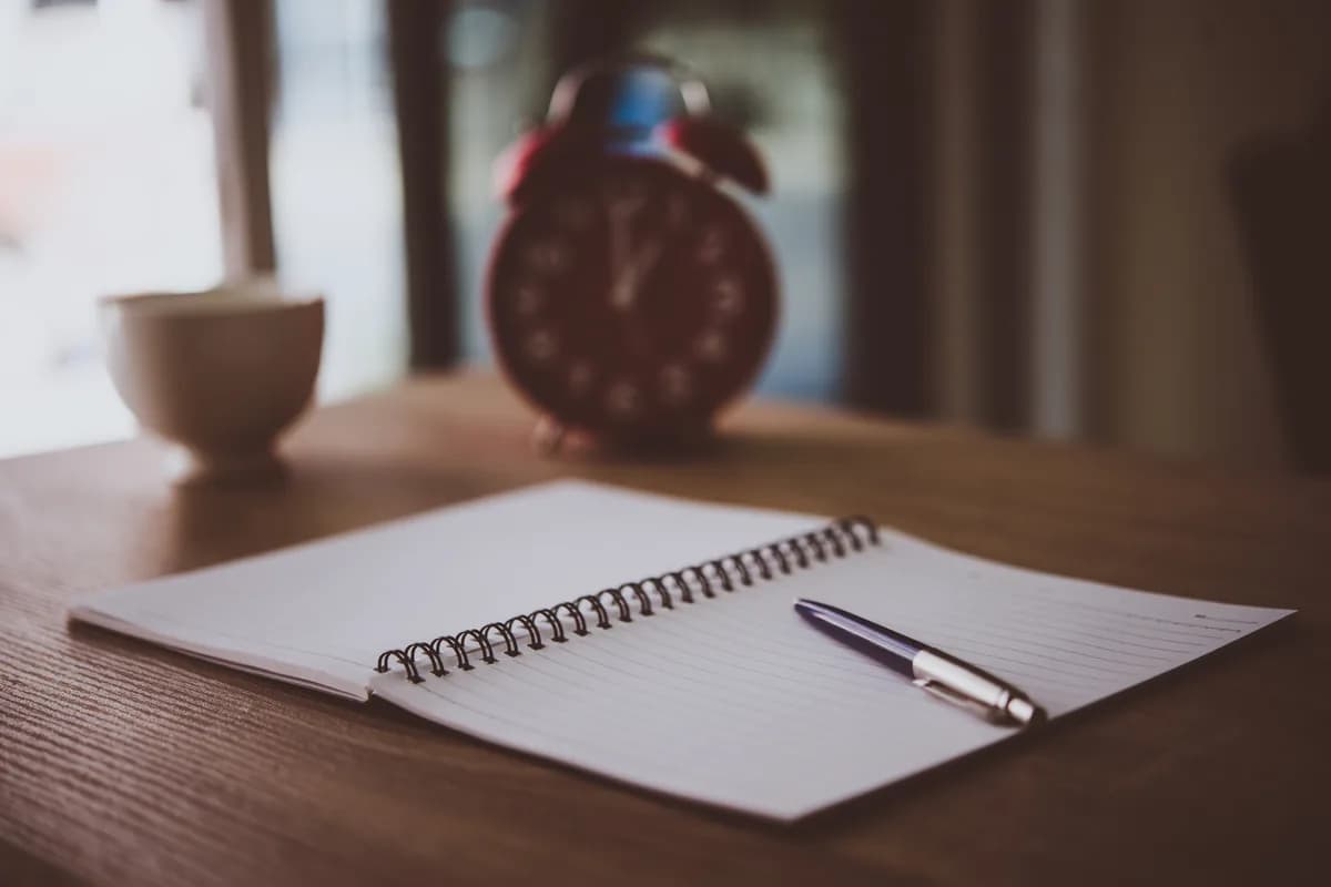Ein Wecker, ein aufgeschlagenes Notizbuch mit Stift und eine Kaffeetasse auf einem Holztisch im warmen Licht
